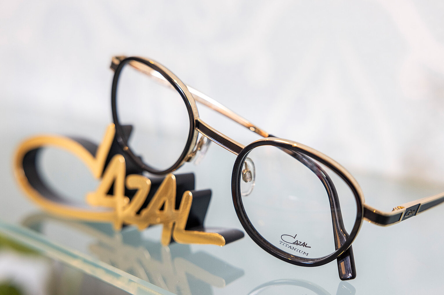Luxusbrille von Cazal im Retrodesign Elegante Brille mit rundem, schwarz-goldenem Rahmen, präsentiert auf Glasfläche neben goldenem Cazal-Logo.