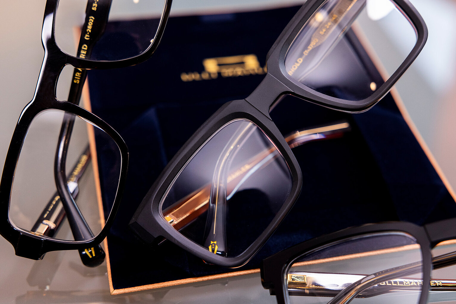 Stilvolle Designerbrillen mit dunkler Fassung Mehrere elegante Korrektionsbrillen mit dunklen Fassungen liegen dekorativ auf einem edlen Etui mit goldenen Details.