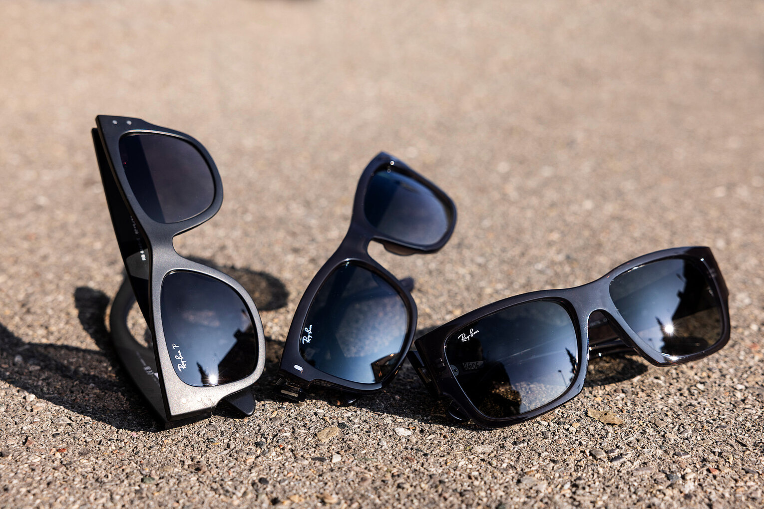 Stylishe Sonnenbrillen für sonnige Tage Drei moderne Sonnenbrillen mit dunklen Gläsern liegen auf einem sonnigen, grobkörnigen Untergrund im Freien.