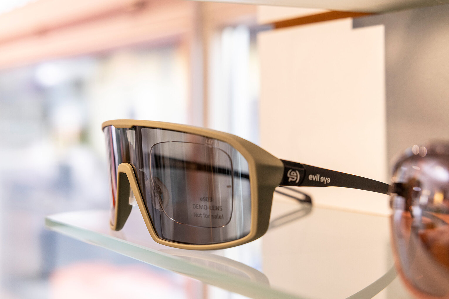 Moderne Sportsonnenbrille mit Panoramaglas Sportliche Sonnenbrille mit breitem, getöntem Glas und markanter Fassung auf einem Glasregal im Optikgeschäft ausgestellt.